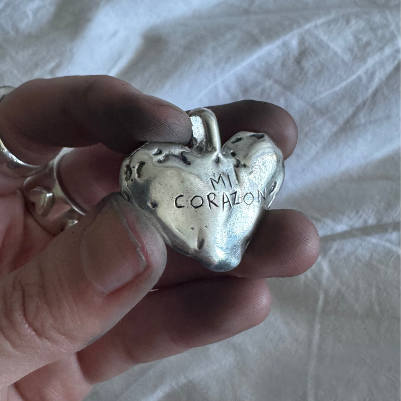 Silver heart-shaped pendant with 'MI CORAZON' engraved, held in a hand against a light background.